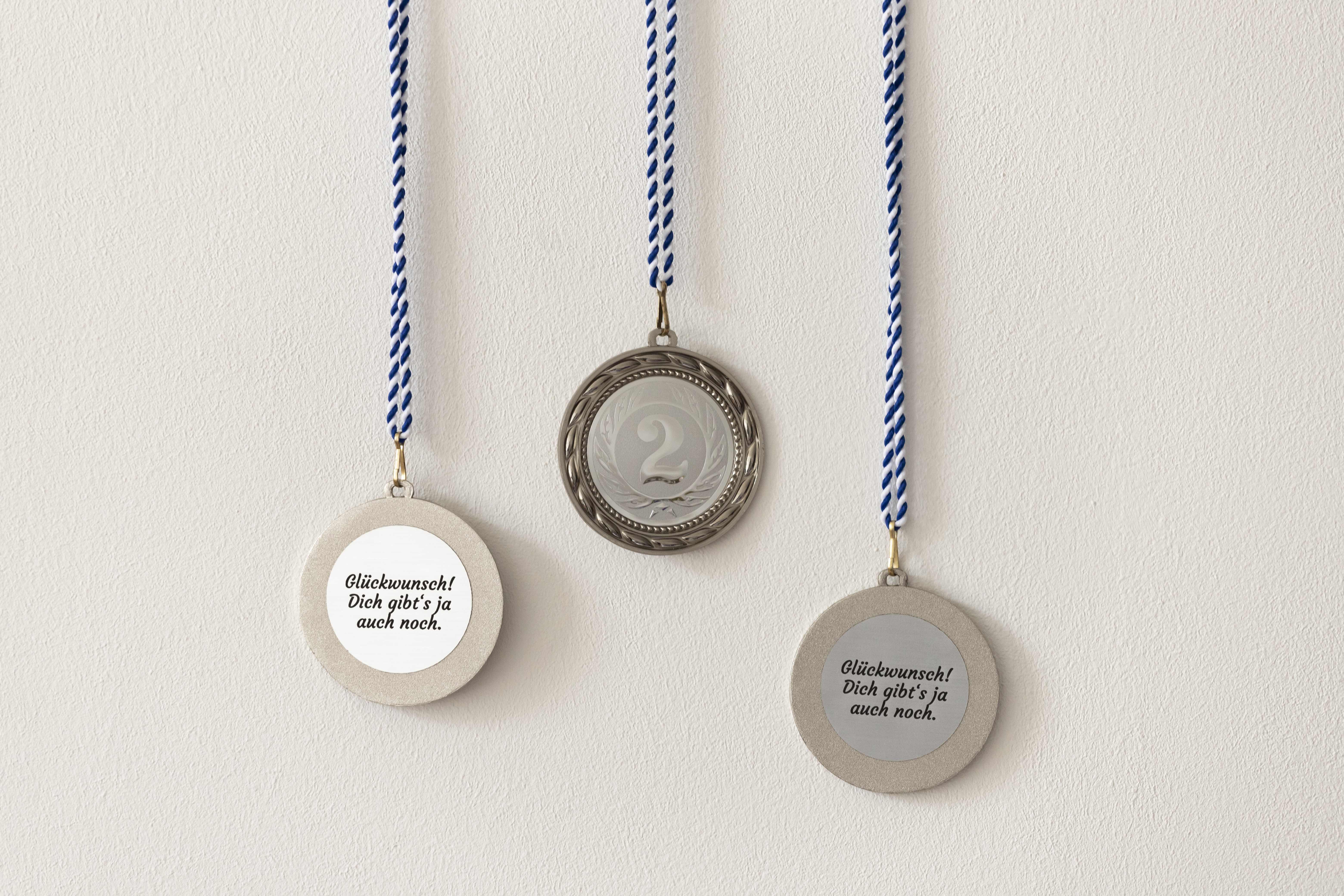 Picture of three silver medals with the number two on the front side of the medal and a sentence on the back  side, that says: Glückwunsch, dich gibt's ja auch noch!